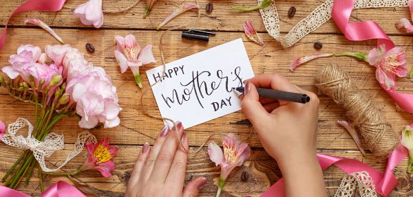 Hands Writing Happy Mother's Day on a card, on top of a wooden table between pink flowers.