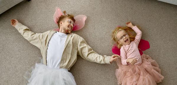 A dad and his daughter both dressed in fairy costumes lying on the floor.