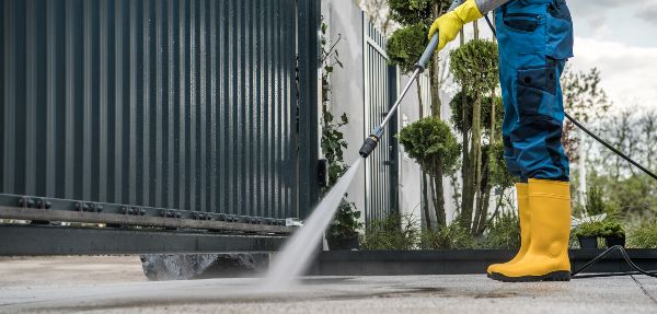 Worker Wearing Protective Uniform and Yellow Rubber Boots Cleaning the Driveway Behind the Entrance Gate Using Pressure Washer Machine. 