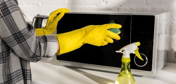 Cropped view of woman in rubber gloves cleaning microwave with sponge.