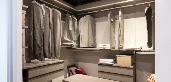 Interior of a walk in closet with hanging clothes and built in drawer units and seating in a luxury modern home.