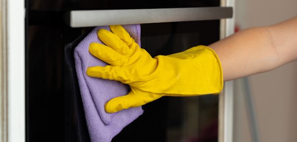 Woman's hand in yellow glove cleaning door of an oven.