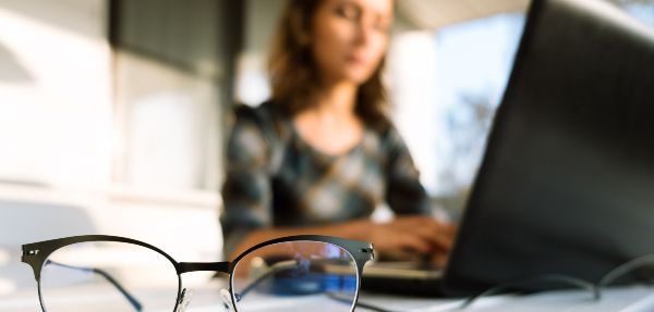 A woman working on a laptop with her glasses in the forefront.