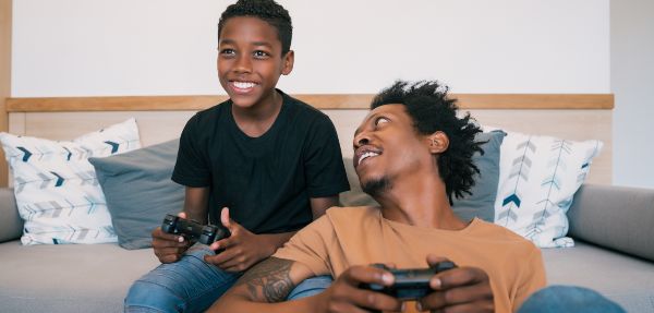 Happy father and son sitting on a sofa couch and playing console video games together at home. 