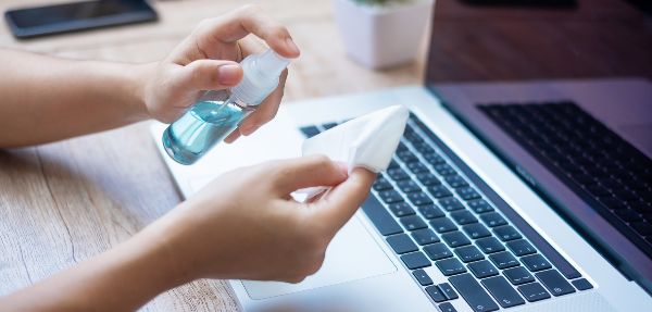 Woman spraying alcohol onto a wet wipe before cleaning a laptop.