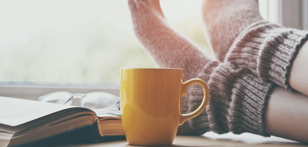 Yellow mug on table in between an open book and a person's feet with socks on