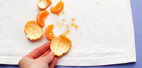 Person's hand holding a naartjie peel beside other peels and naartjie pieces on a stained shirt