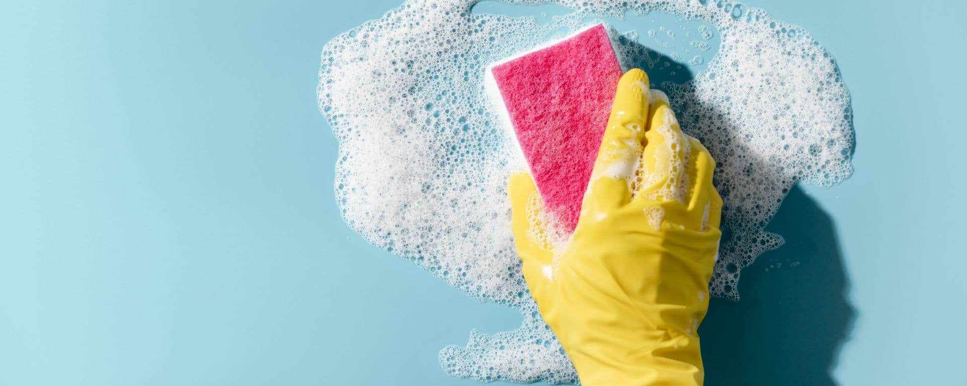 Hand in a yellow rubber glove holds a cleaning sponge and wipes a soapy foam on a blue background.