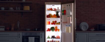 An open fridge full of food with its light on in a dark kitchen